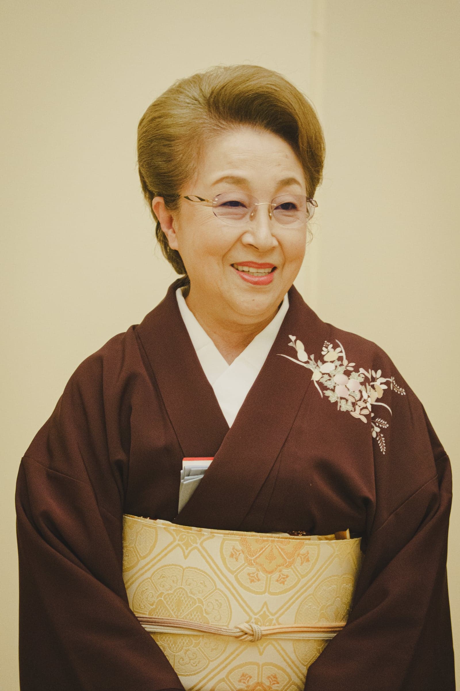 女將千景戴著眼鏡,身穿深紅色和服、繫著金色腰帶,面帶微笑。