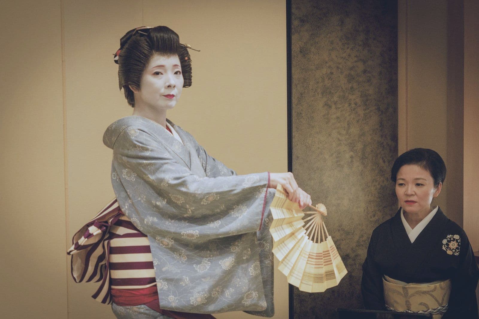 一位穿灰色和服的藝妓手持打開的扇子起舞,另一位藝妓跪坐在旁。