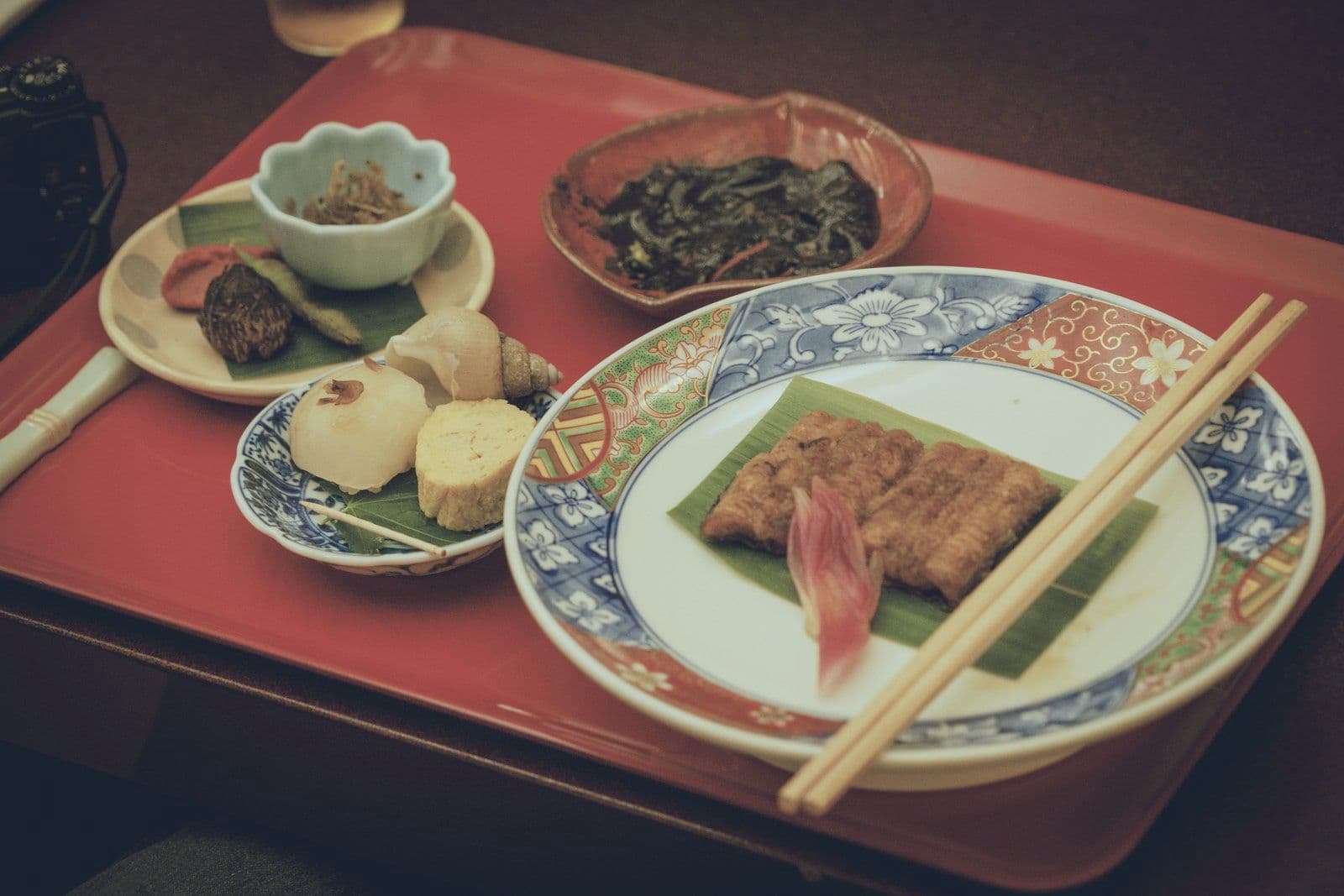 漆盤上擺著放在葉片上的烤鰻魚、幾道小菜和筷子。