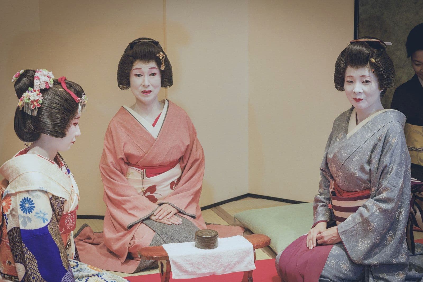 三位穿淡色和服的藝妓並排坐在榻榻米上,對著鏡頭微笑。