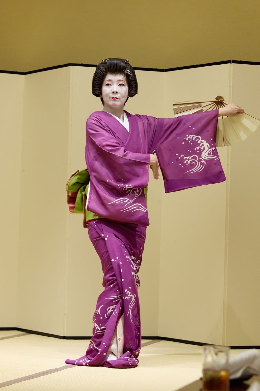 一位穿紫色和服的藝妓,在屏風前持著打開的扇子起舞。